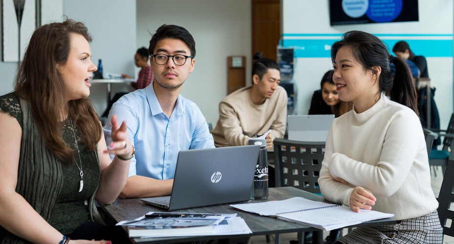 Three students engaged in discussion while working on laptops at a table in a collaborative study environment.