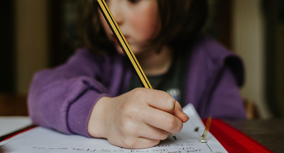 Young girl writing with a pencil in a red book.