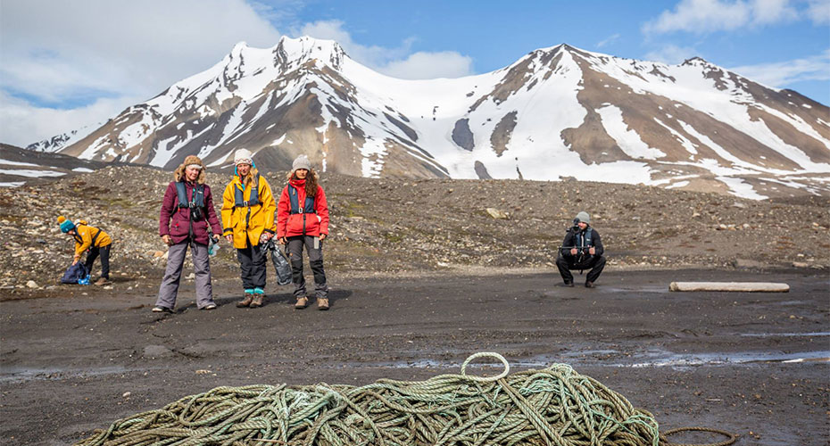 A group of people in outdoor gear standing on a rocky, snow-dotted landscape with mountains in the background. In the foreground, there is a large bundle of rope on the ground. The scene suggests an expedition or environmental cleanup, with participants actively engaged in activities against a backdrop of rugged natural beauty.