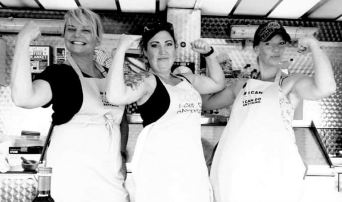 Three women posing and flexing their muscles while smiling. They are wearing aprons that say 