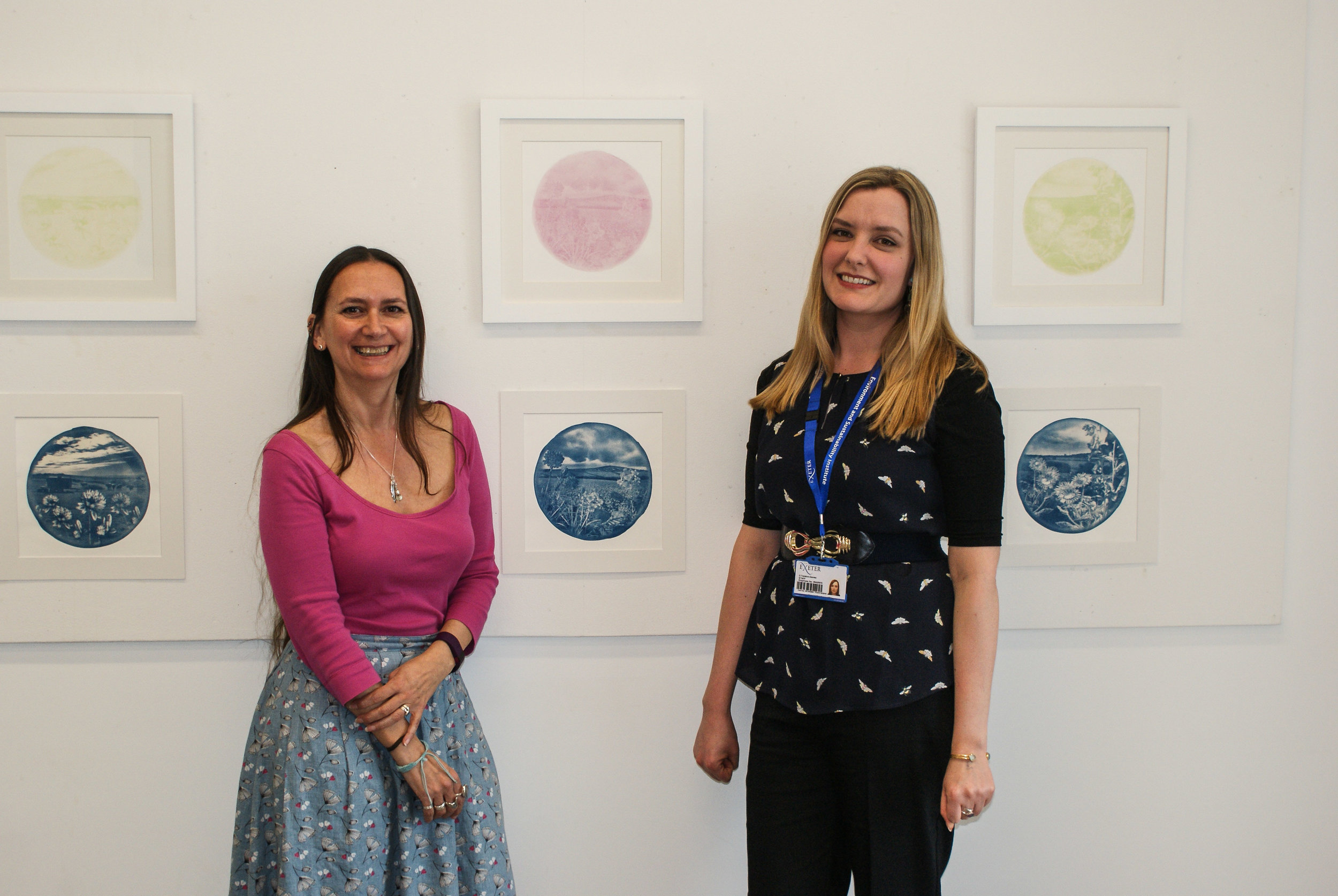 Two women standing in front of a wall displaying framed circular artworks in pastel colors. The woman on the left is wearing a pink top and a patterned skirt, while the woman on the right is wearing a black top with a blue lanyard and dark pants. Both are smiling, creating a welcoming and friendly atmosphere in the gallery setting.