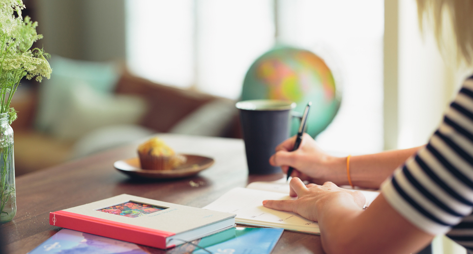 A person sits at a table, focused on writing in her notebook, surrounded by a cozy atmosphere.